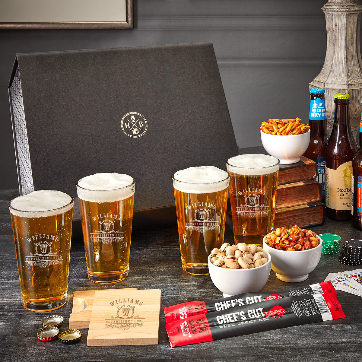 table with a black luxury box, 4 pint glasses full of beer with customization, bowls of peanuts, pretzels, and pistachios, 2 jerky sticks, and a wooden beer-opening coaster